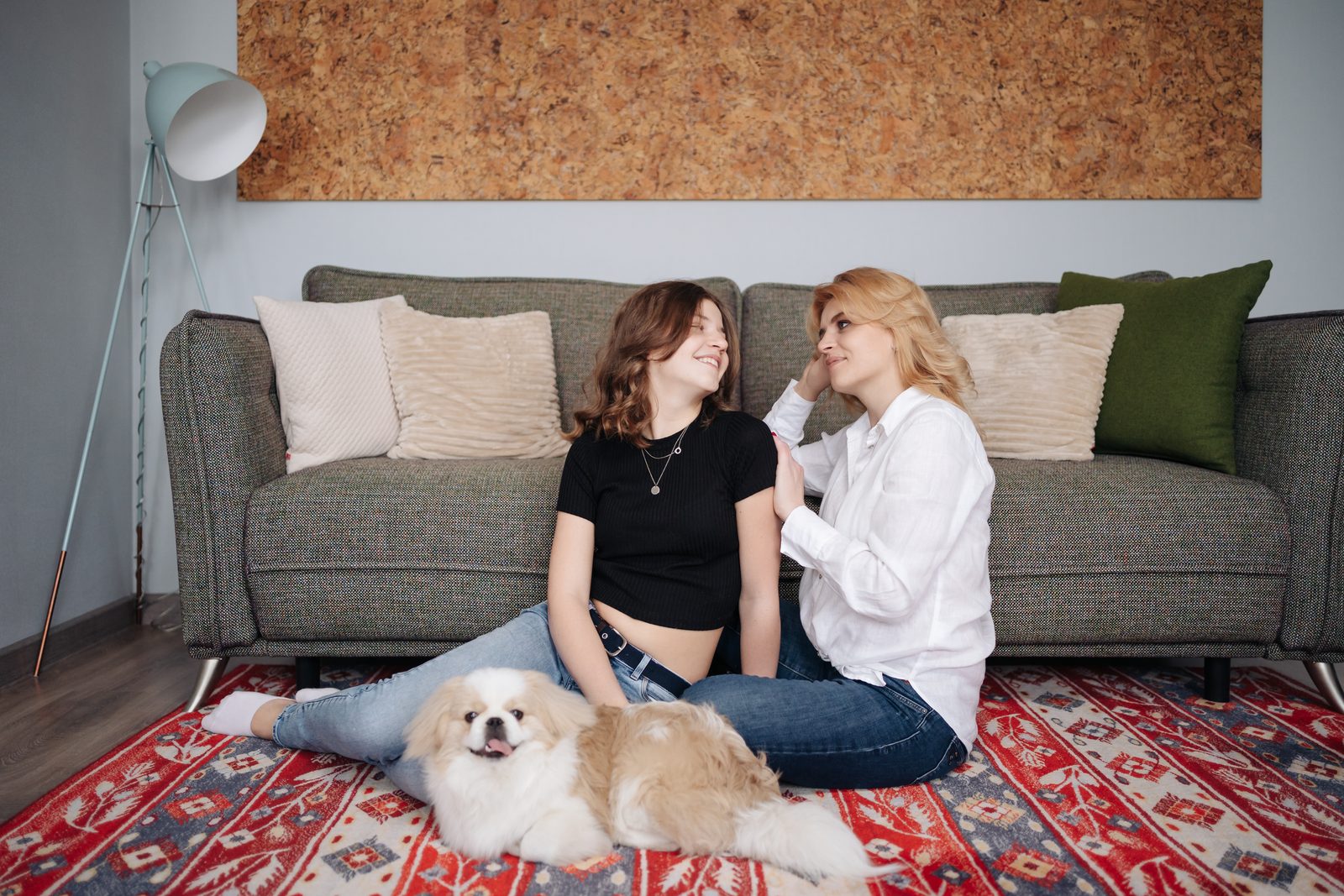 A mother and her adult daughter sit on a red patterned rug against a gray sofa, laughing together beside their small fluffy dog.