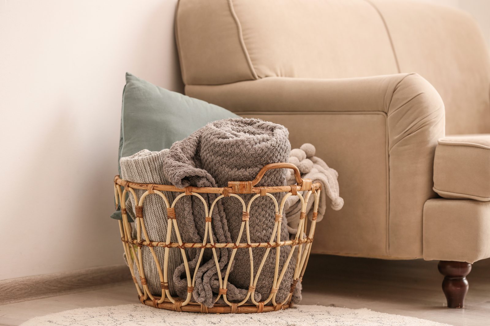 Wicker basket holding folded knit blankets and a green pillow on a rug beside a beige armchair in a bright living room