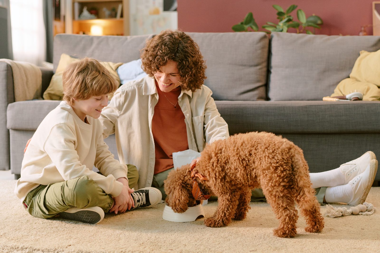 A boy and a woman sit on a living room rug watching a curly brown poodle eat from a white bowl between them