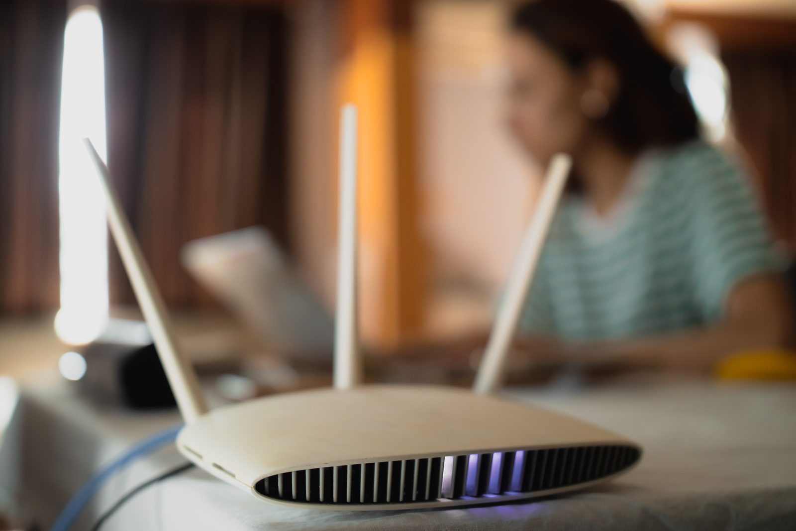Home Wi-Fi router with antennas and a glowing status light on a table, a woman working on a laptop blurred behind it
