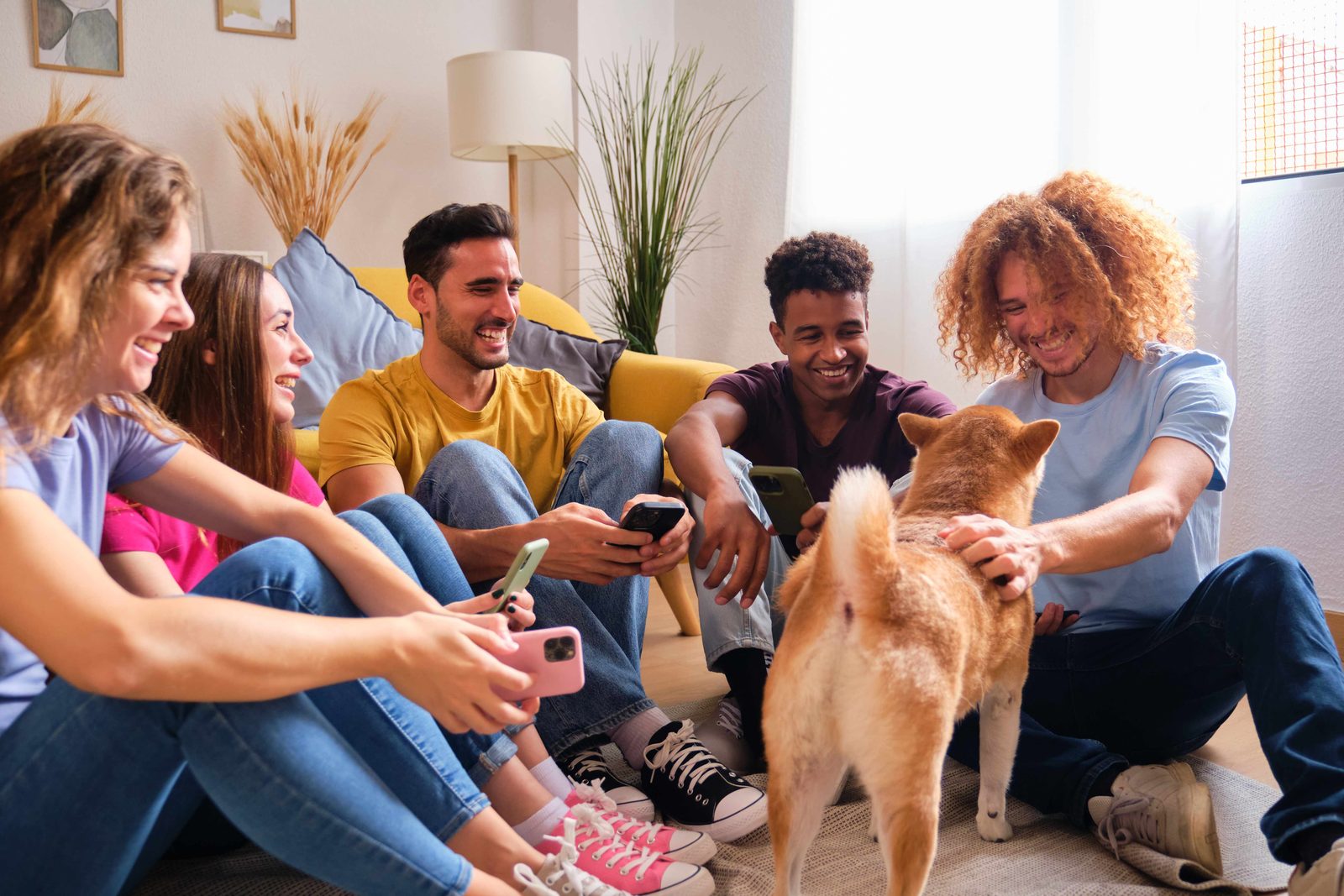 Five young housemates sit together on the living room floor of a shared flat, talking around a Shiba Inu