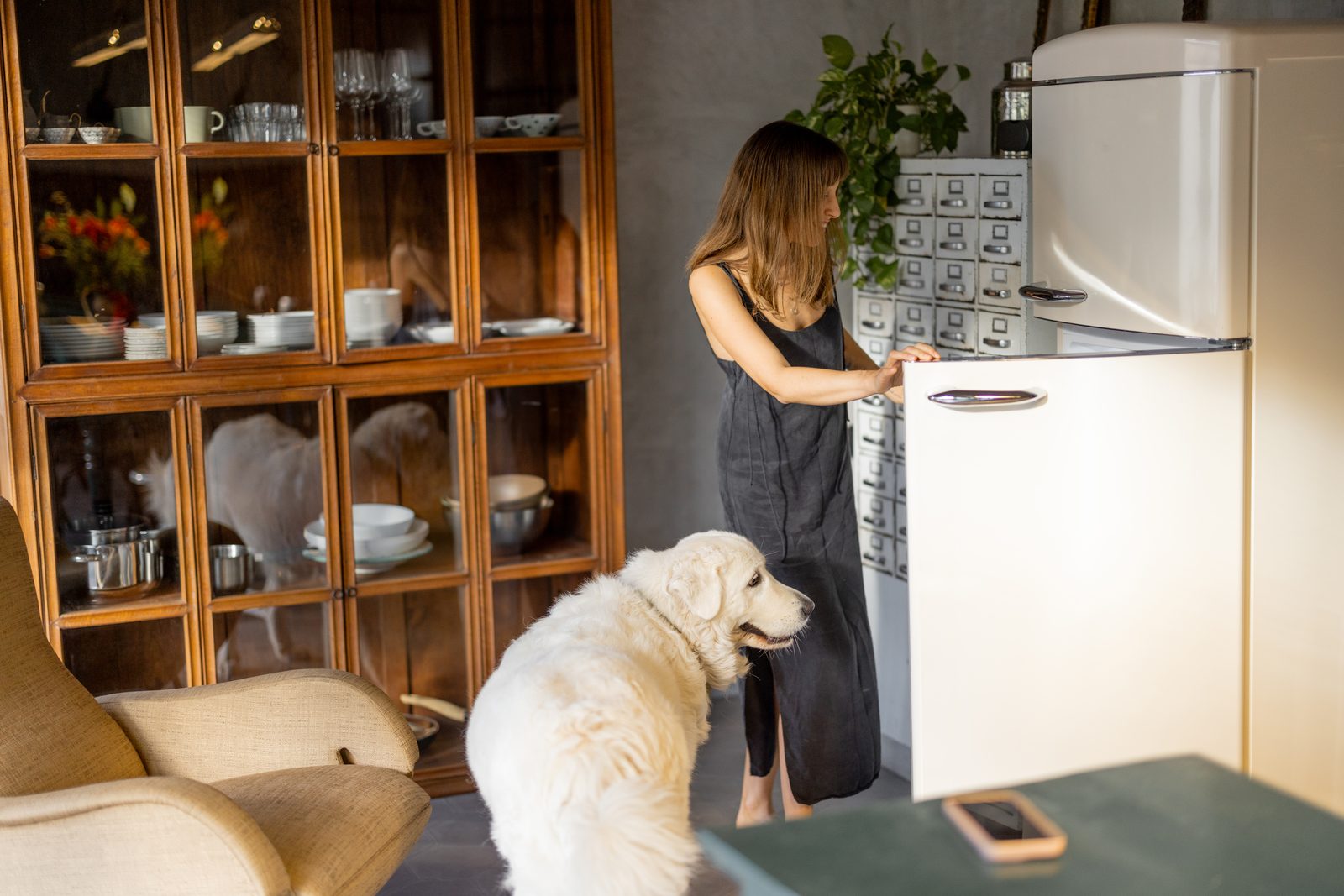Woman opening a cream vintage refrigerator in a warm kitchen while a white fluffy dog stands beside her by a wooden cabinet