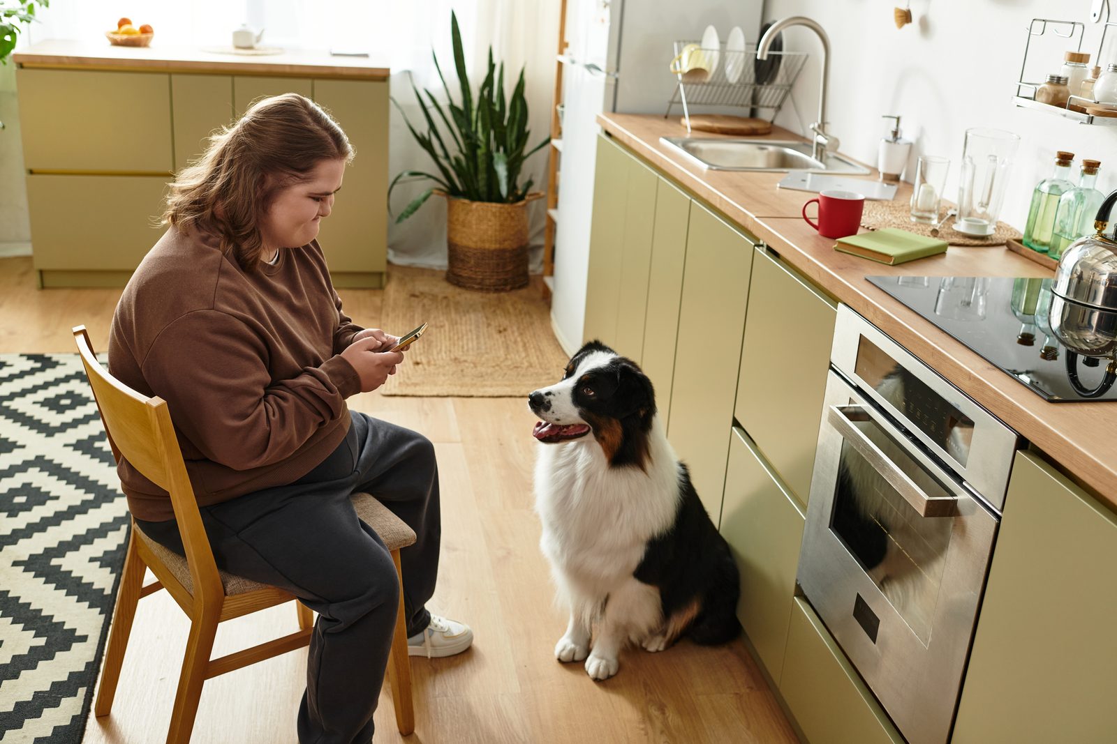 Woman on a wooden chair checking her phone in a green kitchen while an Australian shepherd sits on the floor beside her