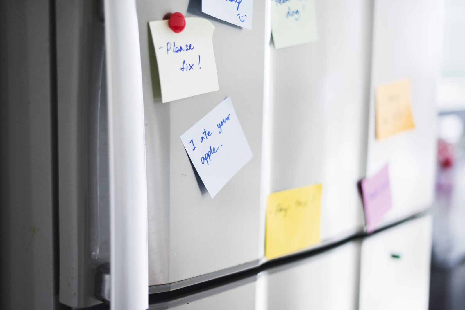 Handwritten sticky notes pinned with a red magnet across a silver refrigerator door in a shared kitchen