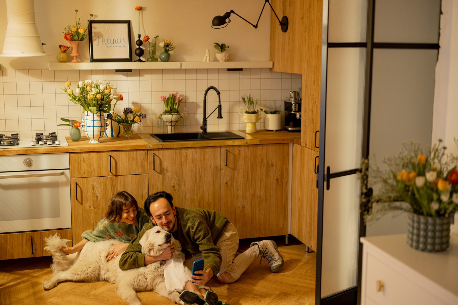 Couple sitting on a warm wooden kitchen floor with a large white dog, the man holding a phone as they talk things over