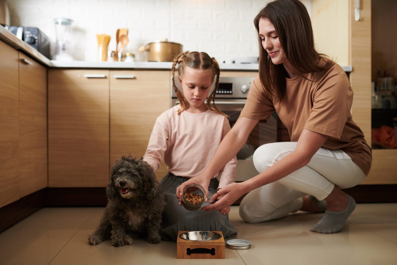 A mother and young daughter kneel on a warm wood kitchen floor, pouring kibble into a bowl beside a curly dark poodle mix.