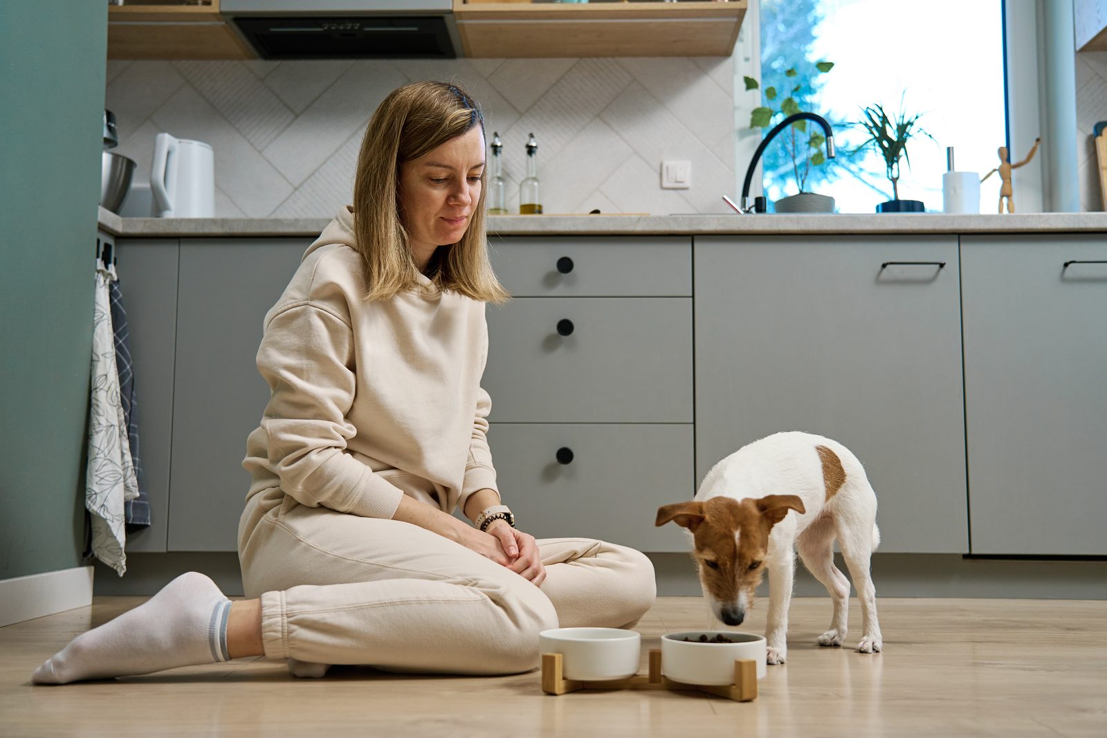 A woman sits on a kitchen floor watching a Jack Russell terrier eat from one of two bowls on a small wooden stand